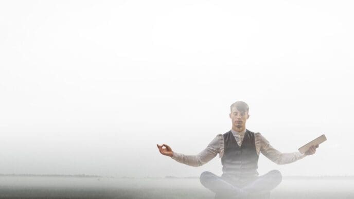 A businessman in a suit sits cross-legged on a yoga mat, meditating in the lotus position.