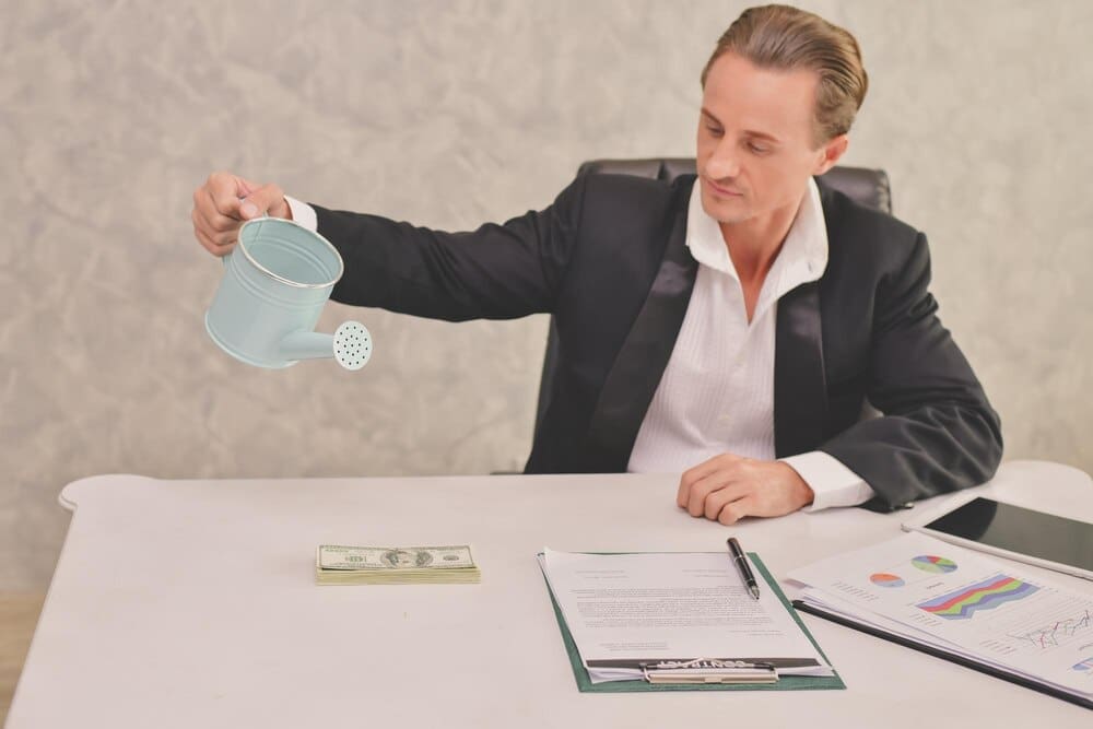 A businessman waters stacks of paper currency with a watering can in an office setting.