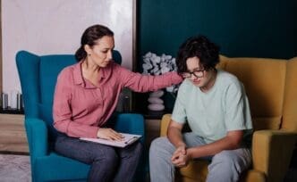 A Caucasian female psychologist consults with a teenage girl, both seated in armchairs in an office setting.
