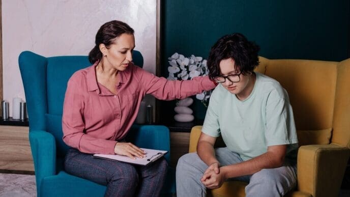 A Caucasian female psychologist consults with a teenage girl, both seated in armchairs in an office setting.
