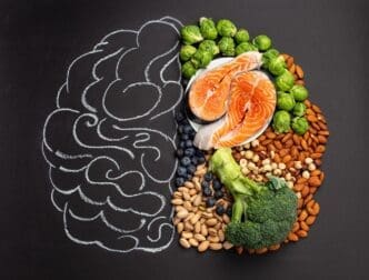 A top-down view showcases a chalk-drawn brain illustration surrounded by various foods like salmon, vegetables, nuts, and berries, symbolizing brain-healthy nutrition against a black backdrop.