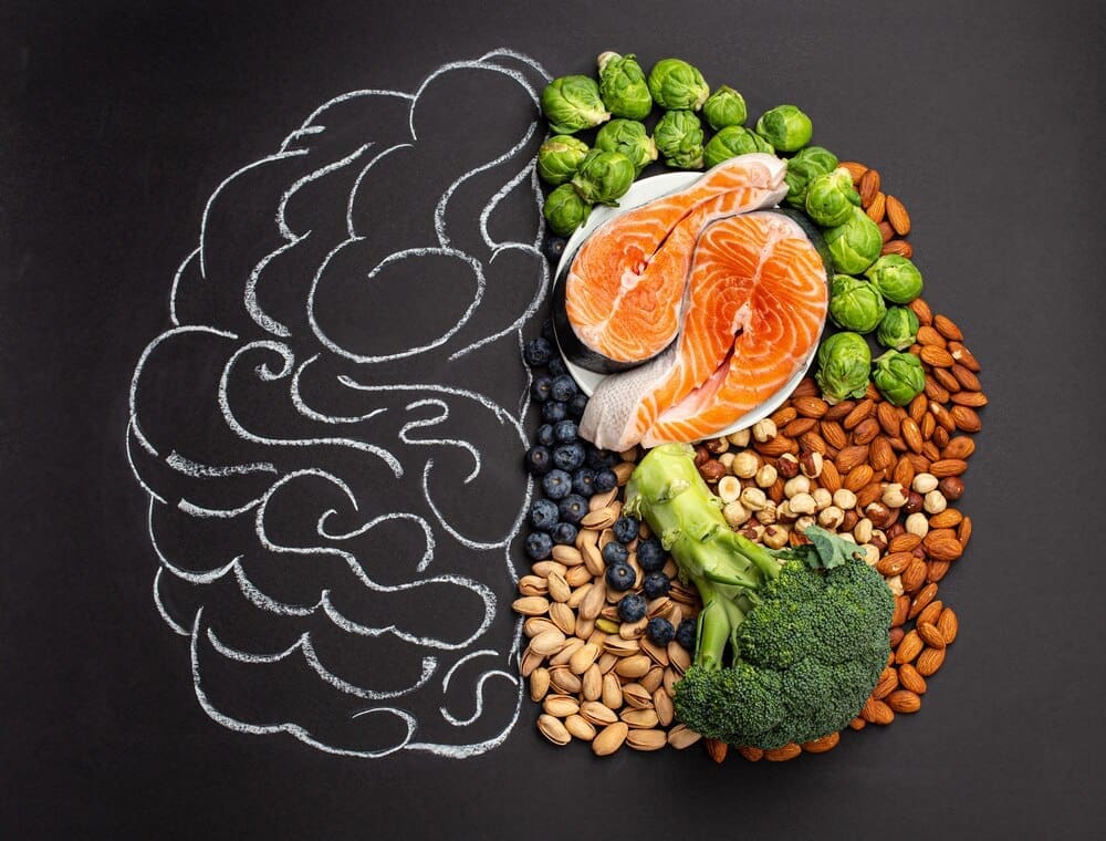 A top-down view showcases a chalk-drawn brain illustration surrounded by various foods like salmon, vegetables, nuts, and berries, symbolizing brain-healthy nutrition against a black backdrop.