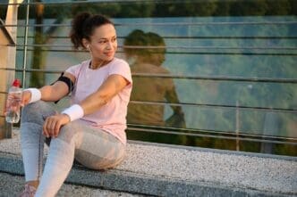 A Black woman in athletic wear rests on a city bridge, holding a water bottle after a morning run at sunrise.