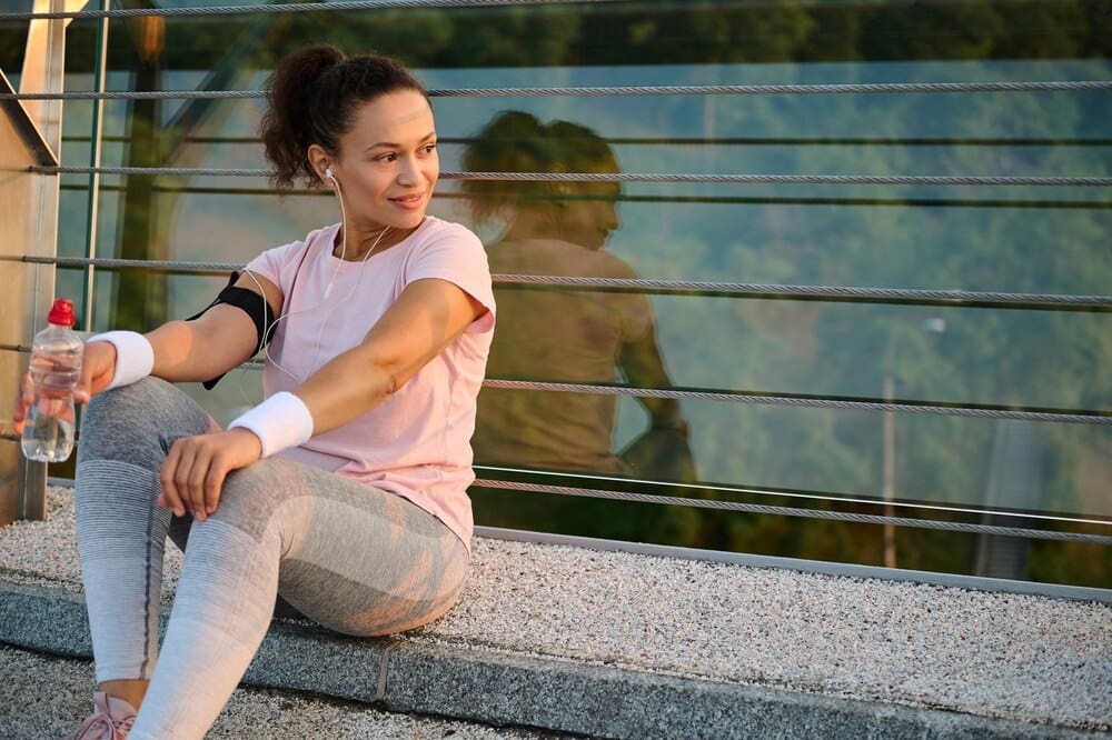A Black woman in athletic wear rests on a city bridge, holding a water bottle after a morning run at sunrise.