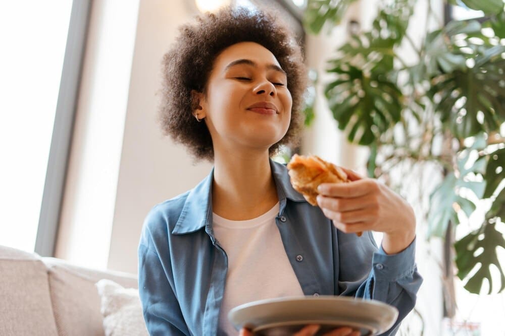 A Black woman with curly hair closes her eyes while enjoying a croissant.