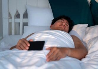 An Asian man with a round face sleeps in a white bed, clutching a smartphone.