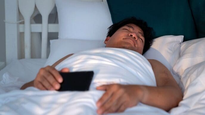 An Asian man with a round face sleeps in a white bed, clutching a smartphone.