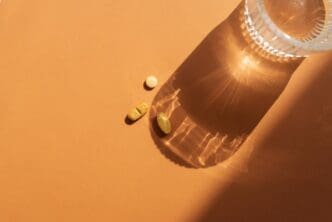 A clear glass of water sits on an orange surface next to several orange pills, casting a shadow.