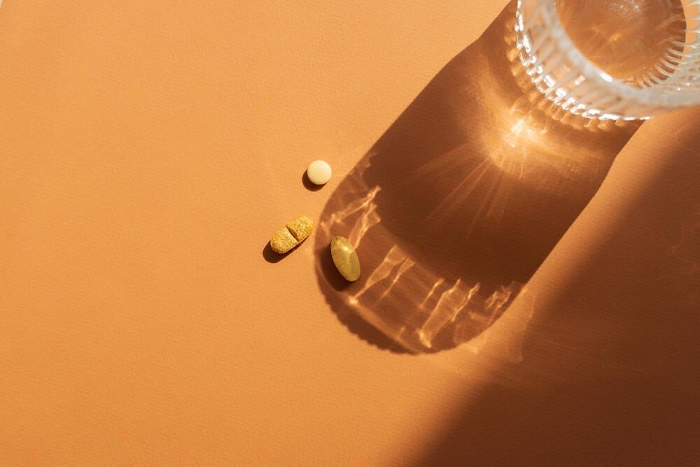A clear glass of water sits on an orange surface next to several orange pills, casting a shadow.