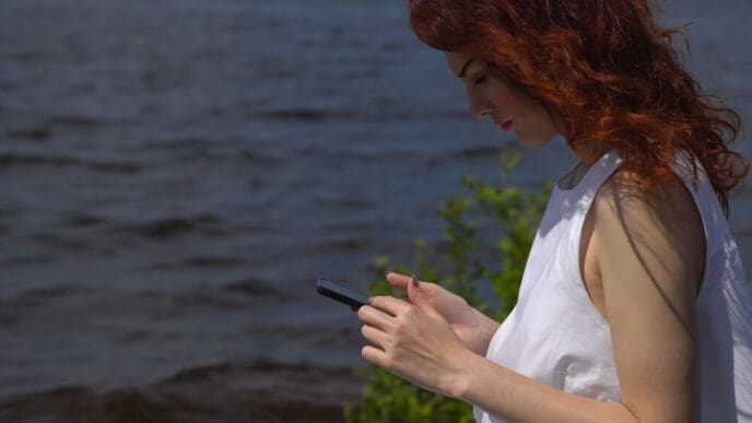 A red-haired girl in a white summer dress uses a cell phone while sitting near a river.