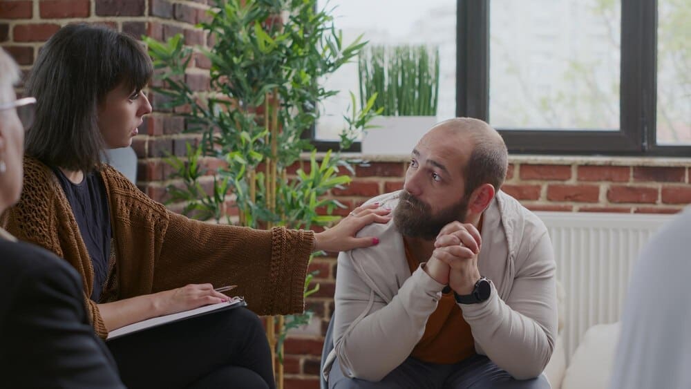 A man trembles and speaks in a group therapy session, discussing anger management with others seated in a circle.