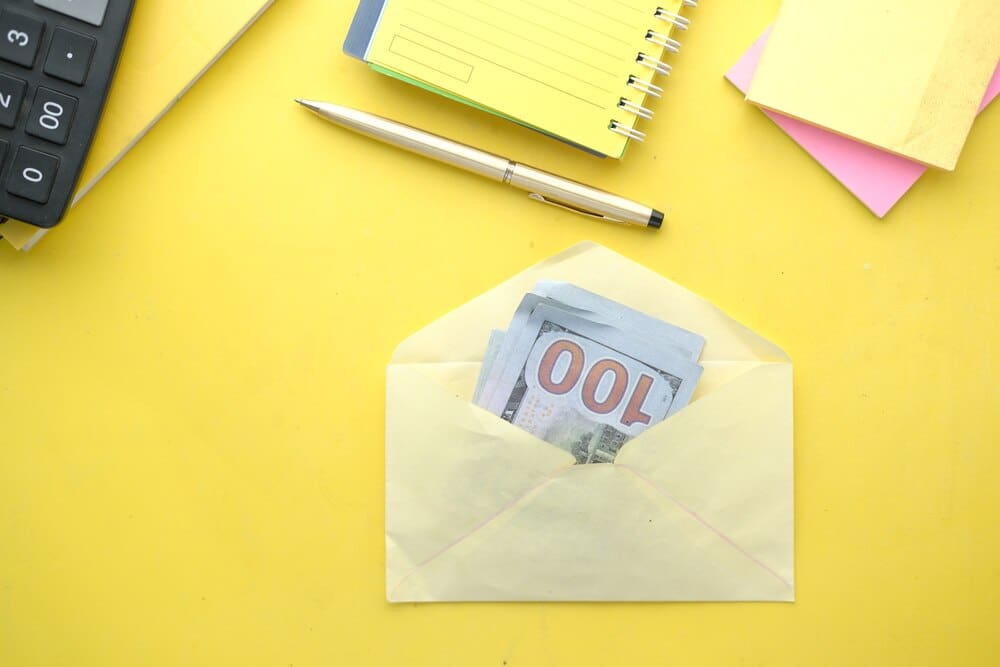 Close-up of a white envelope on a yellow surface, containing a stack of U.S. dollar bills.