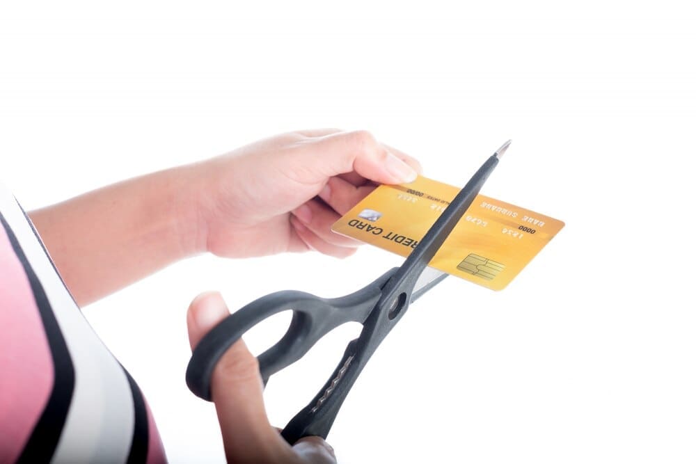 Close-up of a young woman's hand cutting a credit card with scissors against a pink background.