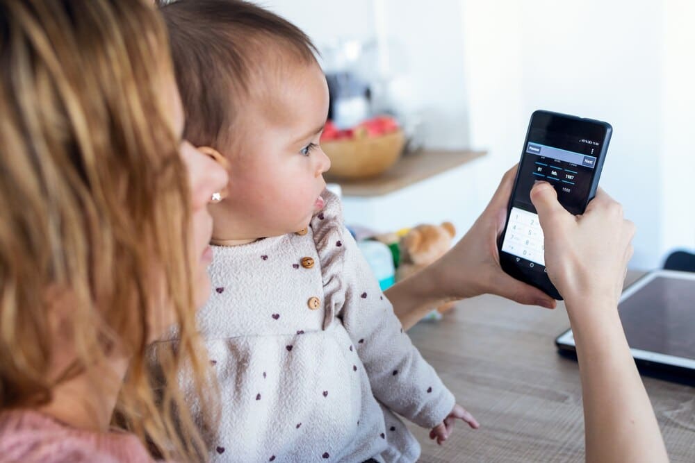 Close-up of a young woman and her baby girl at home, with the woman using a mobile phone and the baby looking at it.