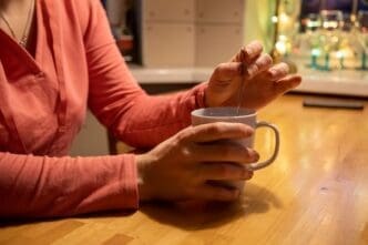 Close-up of a person's hands holding a white mug with a warm beverage, stirring with a teaspoon on a wooden table.