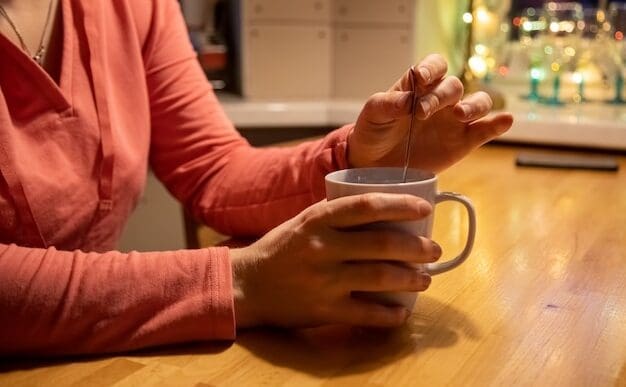 Close-up of a person's hands holding a white mug with a warm beverage, stirring with a teaspoon on a wooden table.