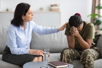 A psychologist, who appears to be White, gently comforts an African American military woman in camouflage fatigues who is sitting on a sofa with her head in her hands, during a therapy session.