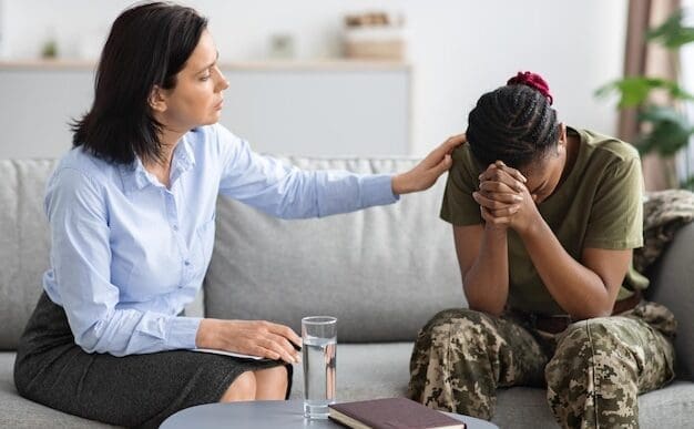 A psychologist, who appears to be White, gently comforts an African American military woman in camouflage fatigues who is sitting on a sofa with her head in her hands, during a therapy session.