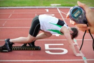 A hand holding a timer is positioned in front of an athletic man at the starting line of a race.