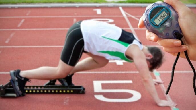 A hand holding a timer is positioned in front of an athletic man at the starting line of a race.