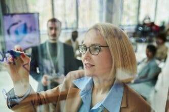 A mature businesswoman with blond hair, wearing a blazer, writes notes on a transparent board.