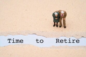 Miniature figures of an elderly couple stand next to the word "retire" on a brown paper background, representing retirement planning.
