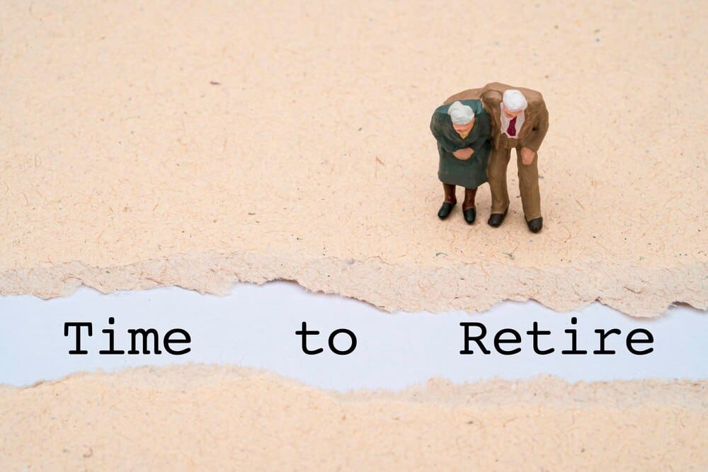 Miniature figures of an elderly couple stand next to the word "retire" on a brown paper background, representing retirement planning.