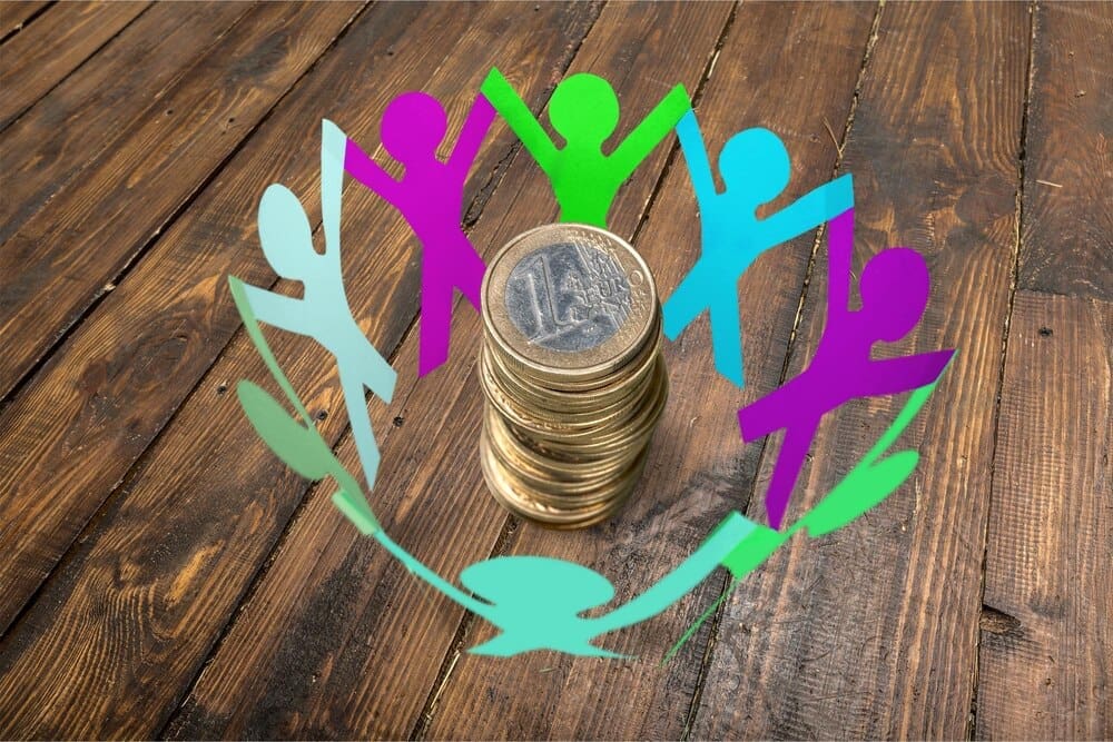 Paper cut-out figures gather around a stack of hundred-dollar bills, illustrating a crowdfunding concept.