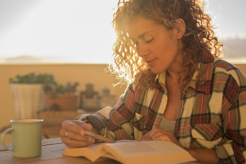 A woman with curly blonde hair reads a book while sitting at a table on a terrace with a sunny sky.
