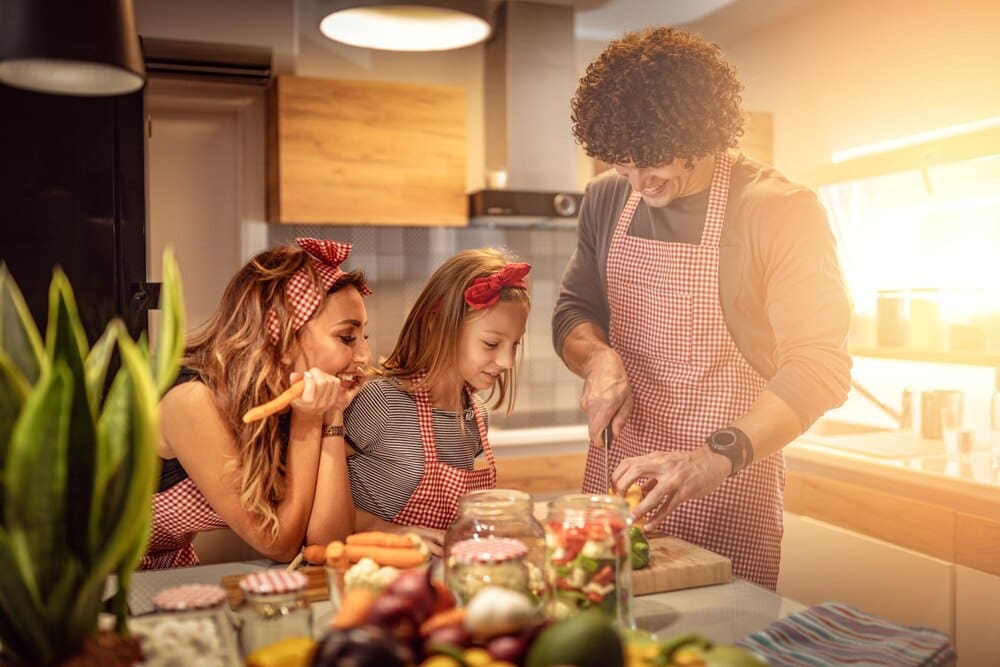 A young girl smiles while cutting vegetables with her parents in a kitchen.