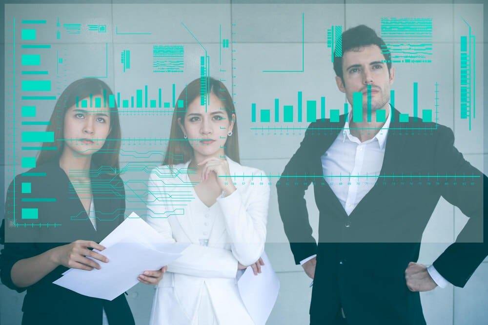 Business colleagues stand together in an office, gesturing toward a projected image on an invisible screen.