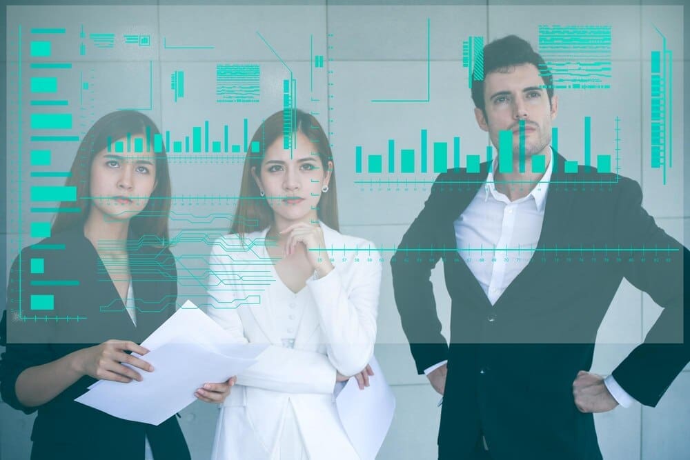 Business colleagues stand together in an office, gesturing toward a projected image on an invisible screen.