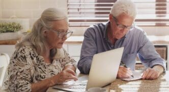 An elderly couple reviews financial documents and uses a laptop in their kitchen, likely for budgeting and online banking.