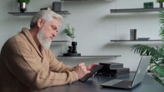 An elderly man with a gray beard draws on a tablet in his living room.