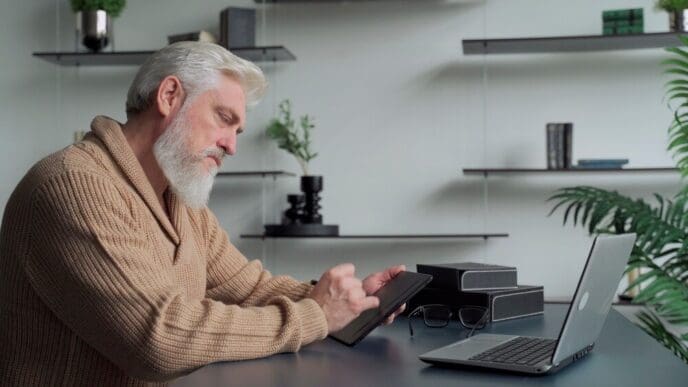 An elderly man with a gray beard draws on a tablet in his living room.