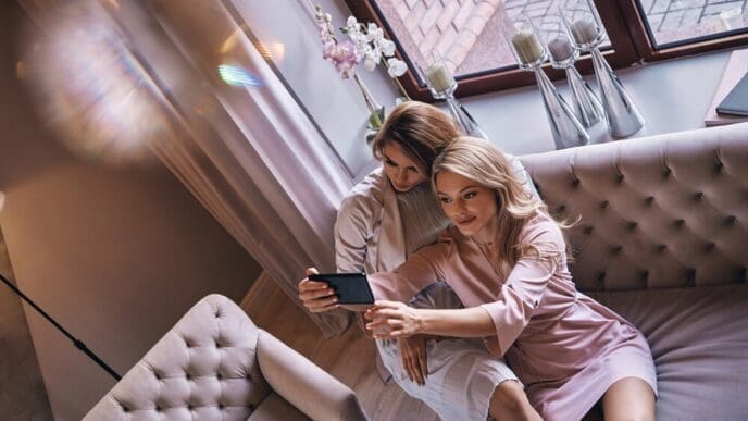 Top-down view of smiling young women in elegant dresses taking a selfie on a sofa.