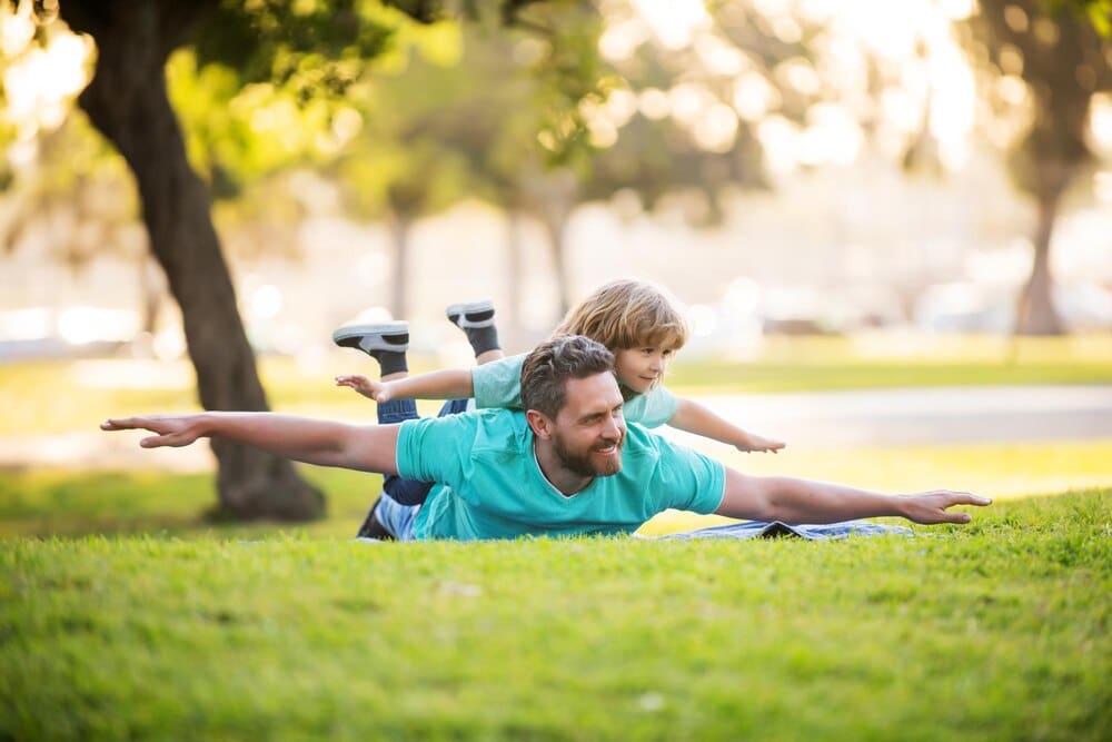 A father lies on the grass, holding his excited young son on his shoulders, celebrating a weekend together.