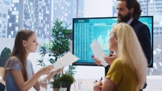 Financial advisor pointing at a large screen displaying financial graphs and data to a group of customers.