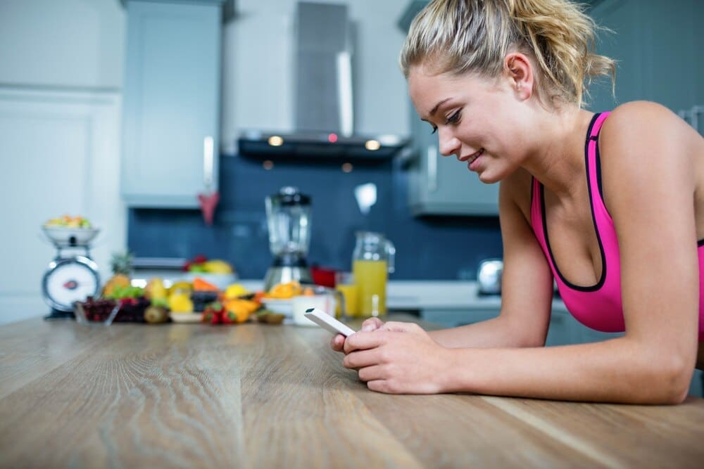 A young woman in workout clothes stands in a kitchen, smiling as she types on her phone.
