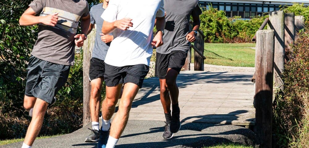 Four cross-country runners in motion, making a left turn while crossing a wooden bridge on a path.