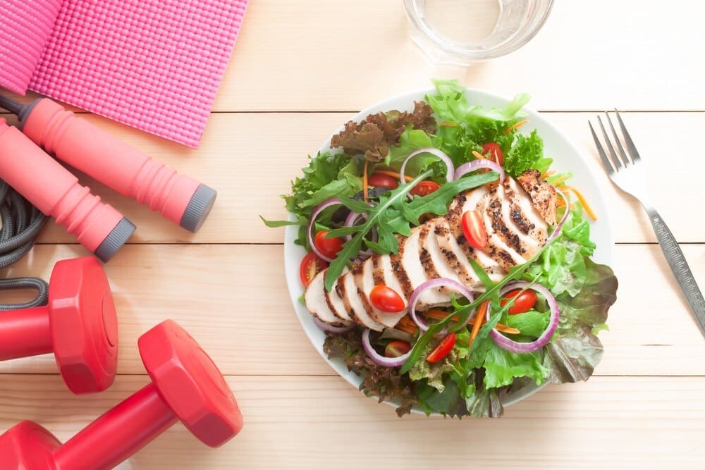 A fresh salad with chicken sits on a wooden table next to fitness equipment.