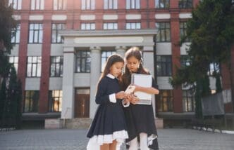 Two young schoolgirls stand side-by-side outdoors near a school building.