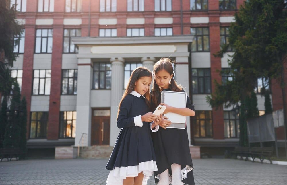 Two young schoolgirls stand side-by-side outdoors near a school building.