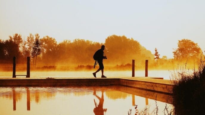 A man walks on a pier extending into a lake, silhouetted against the sunrise.