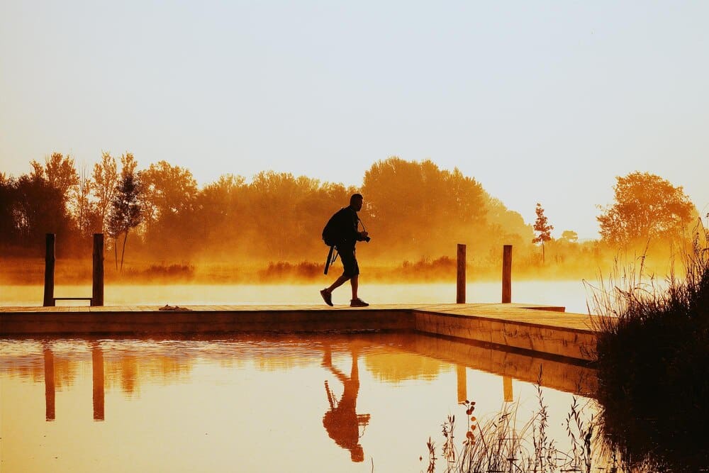 A man walks on a pier extending into a lake, silhouetted against the sunrise.