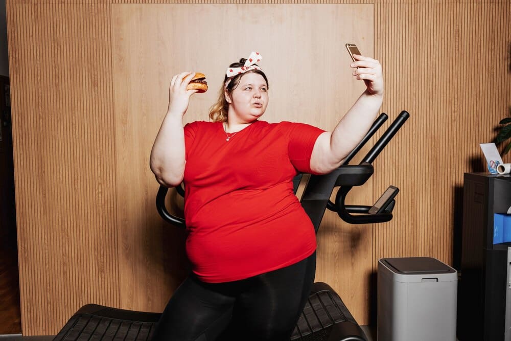 A woman in workout clothes and a bandage poses for a selfie with a burger next to a treadmill.