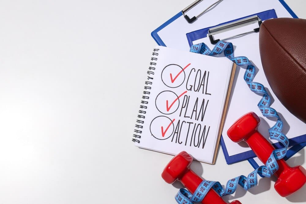 Notebook open to a goal plan with a pair of dumbbells and a rugby ball nearby.