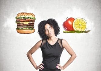 A young African American woman in black jeans stands with her hands on her waist, contemplating near a concrete wall with hamburger and fruit sketches.