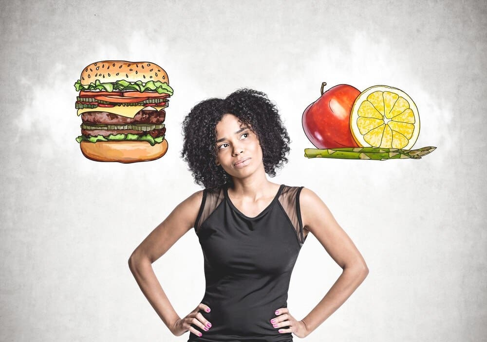 A young African American woman in black jeans stands with her hands on her waist, contemplating near a concrete wall with hamburger and fruit sketches.