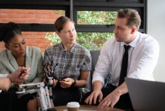 A diverse group of businesspeople brainstorm around a hand-shaped robot model in an office setting.